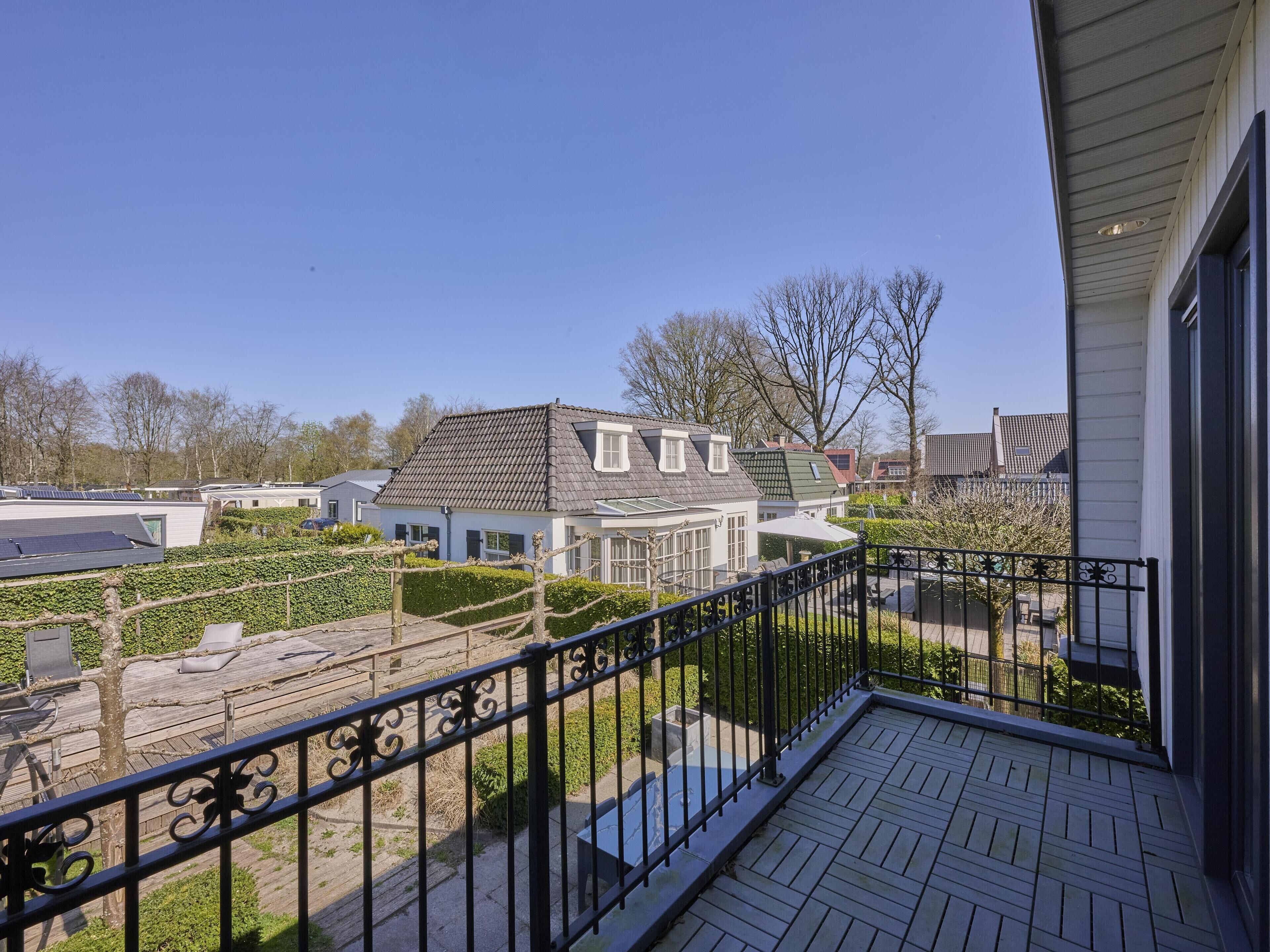 Een woongebied met een huis, een schuin dak, een balkon met een zwarte metalen balustrade en uitzicht op omliggende huizen en bomen.