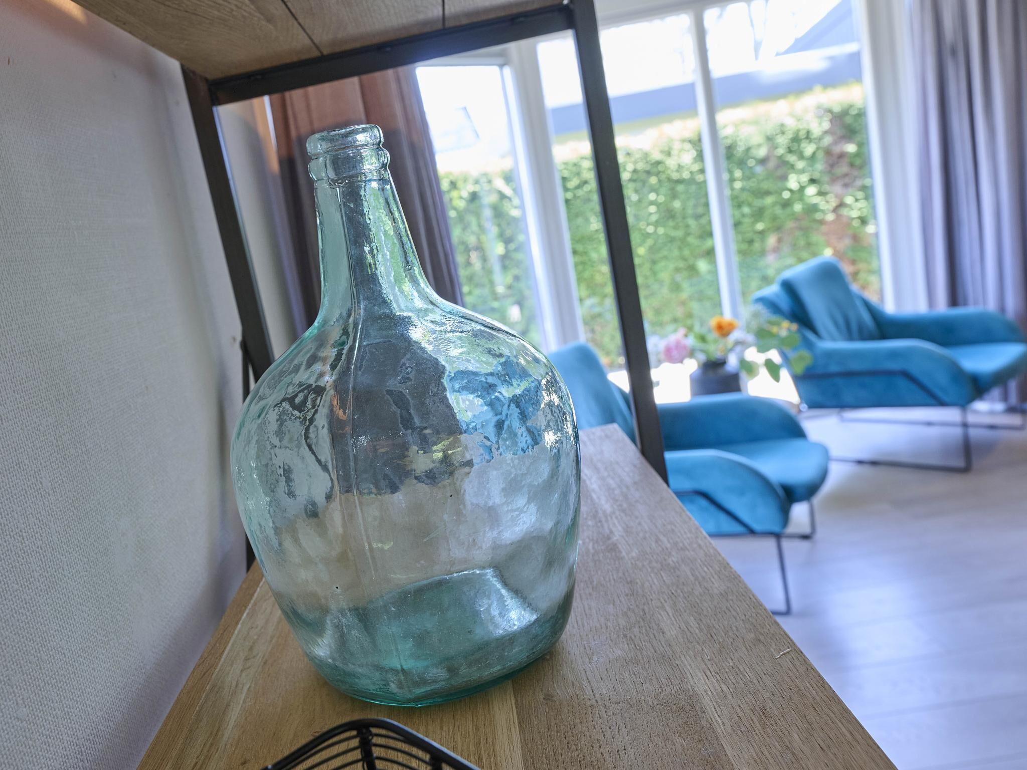 En genomskinlig glasflaska står på ett träbord i ett rum med modern inredning, stora fönster och blå möbler.