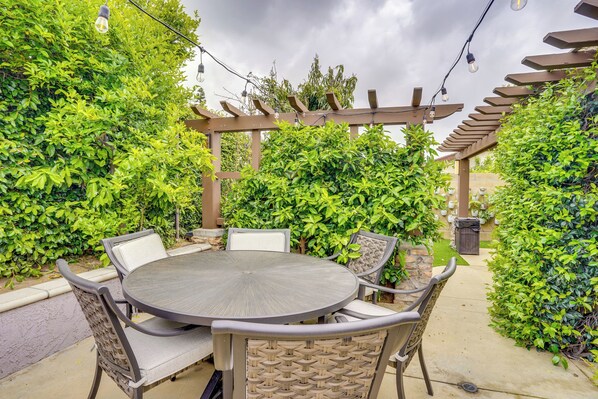 Un patio exterior con una mesa redonda y sillas, rodeado de exuberante vegetación y una pérgola.