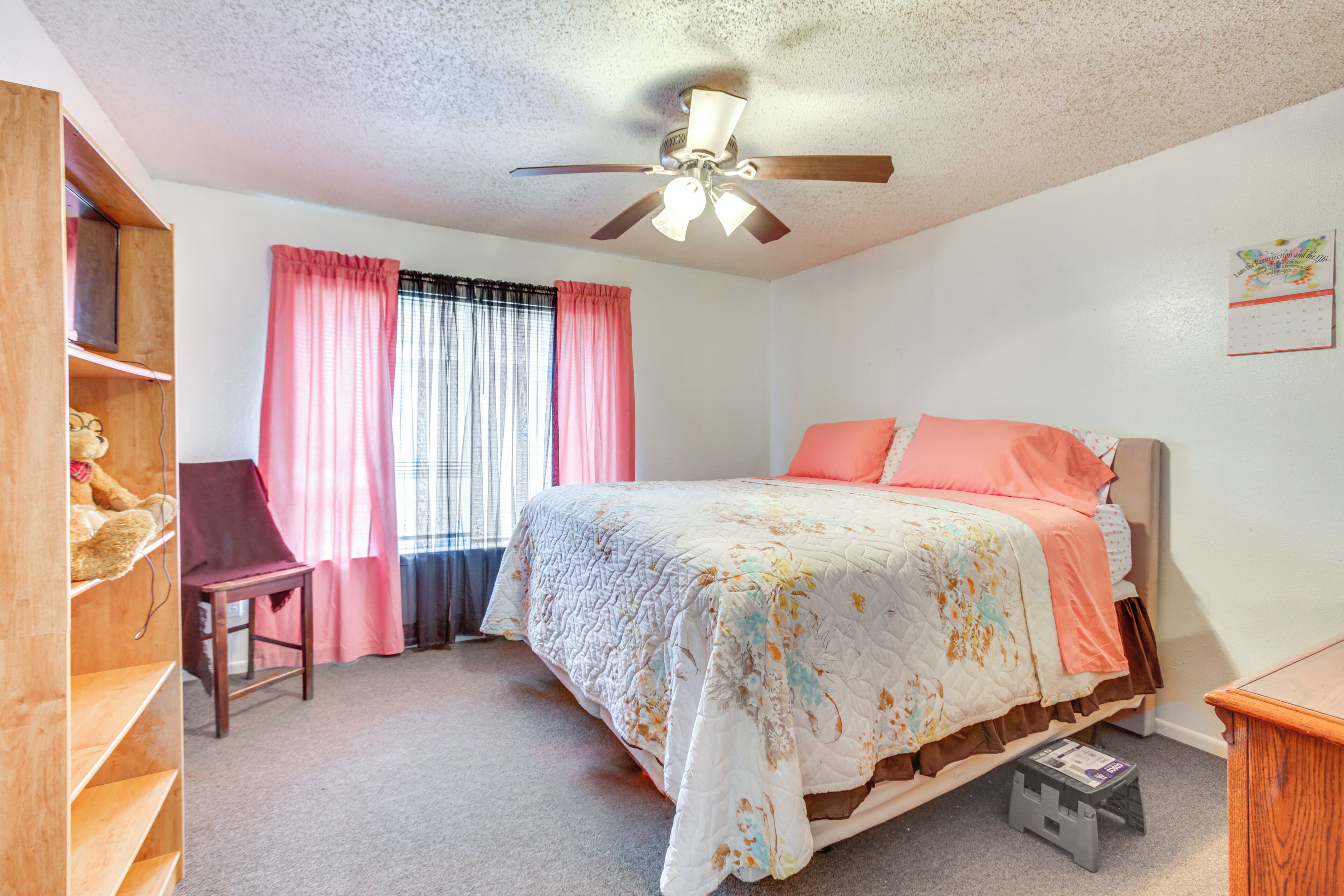 A bedroom with a bed, a desk, a chair, a ceiling fan, and a calendar on the wall.