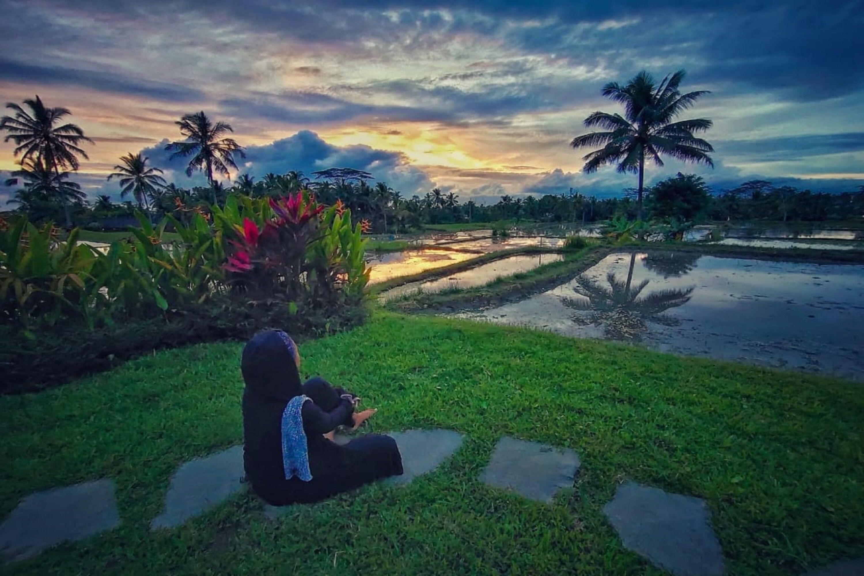 A person sitting on a stone path in a garden with palm trees and water features.