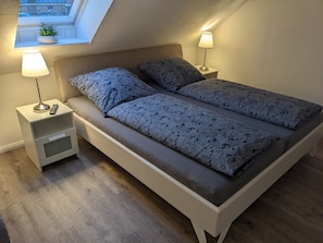 Une chambre avec un plafond en pente, un lit avec une literie à motifs, des lampes de chevet, une petite table de nuit et une fenêtre avec une plante.