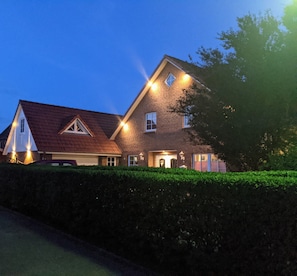 Une grande maison à deux étages, dotée d’un toit en pente, de nombreuses fenêtres et d’une haie bien entretenue à l’avant.