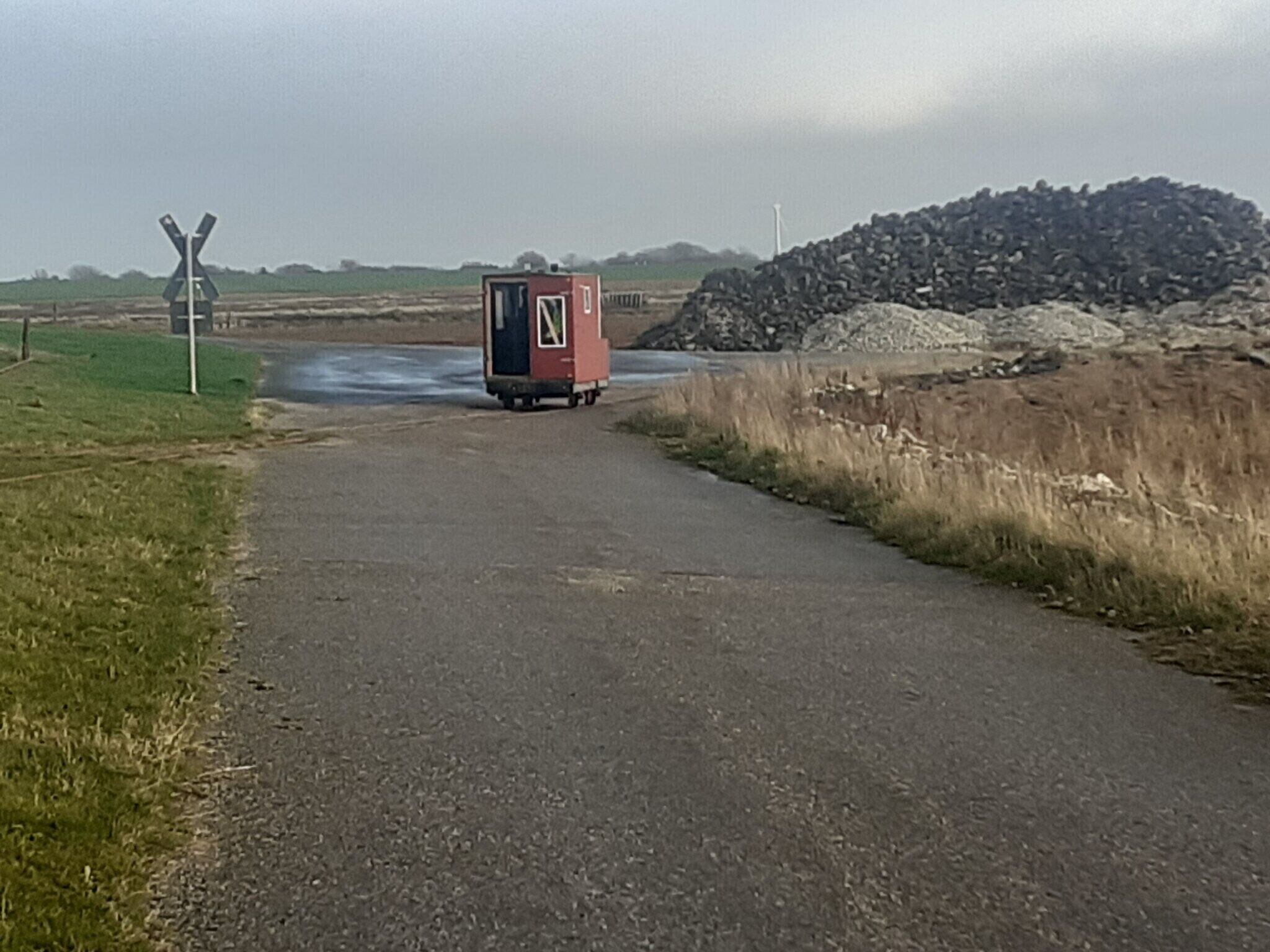 Een landelijke weg met een rode mobiele toiletten en een spoorwegovergang.