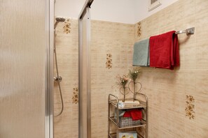 Un espace douche doté d’une porte en verre, d’un porte-serviettes avec plusieurs serviettes et d’une petite étagère sur laquelle sont posés un vase et des objets décoratifs.