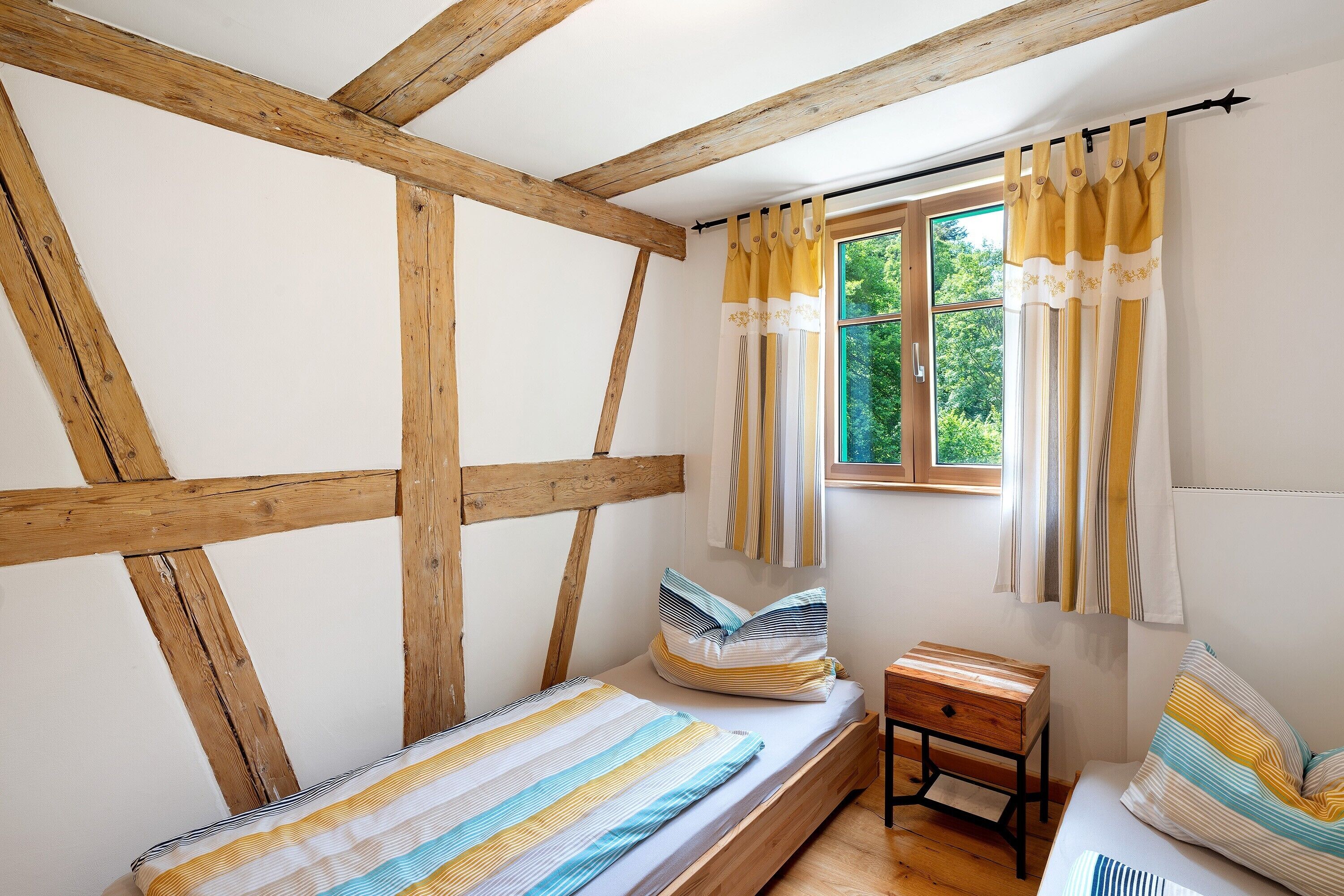 Une chambre avec un plafond orné de poutres en bois, un lit simple avec une literie à rayures, une petite table de chevet et une fenêtre avec des rideaux.