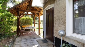 A covered patio with a wooden pergola, a table with chairs, and a door leading to a building.