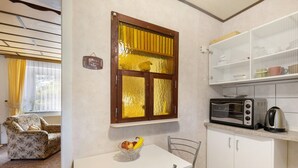 A compact kitchen with a microwave, toaster, and kettle, a dining table with chairs, a floral-patterned sofa, and a window with yellow curtains.