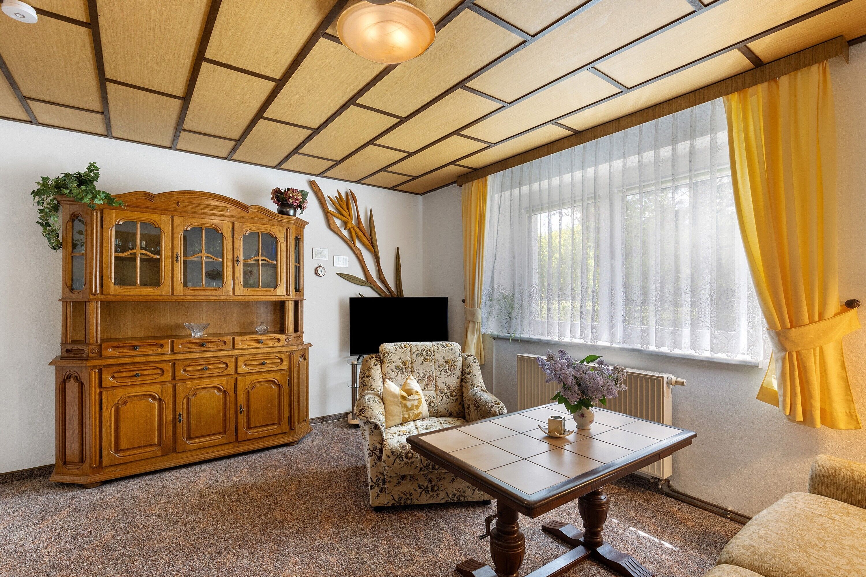 Uma sala de estar com um armário de madeira, uma poltrona estampada, uma mesa e uma grande janela com cortinas amarelas.