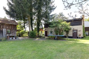 A rustic wooden building with a large wheel, a modern house with a brown door, and a grassy yard.