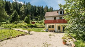 Une maison avec un balcon rouge, entourée d’un jardin et d’une forêt.
