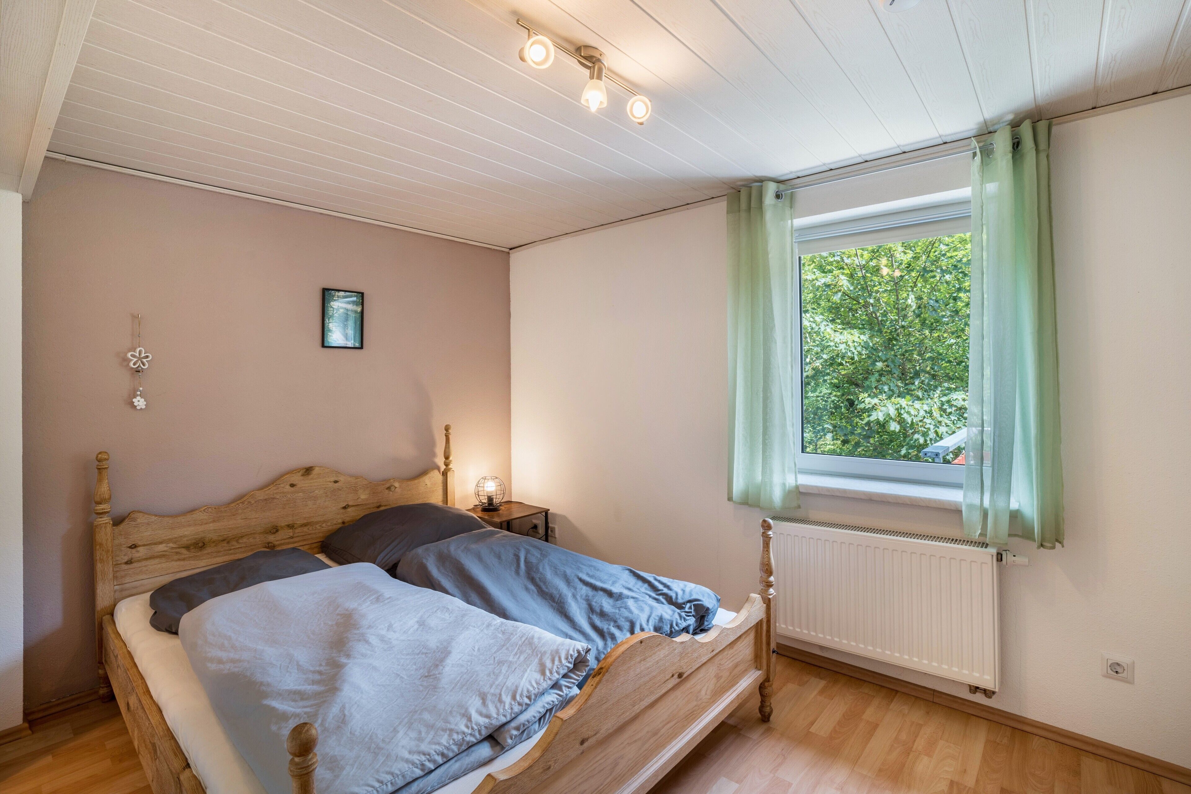 A bedroom with a wooden bed, a bedside table with a lamp, a window with curtains, and a radiator.