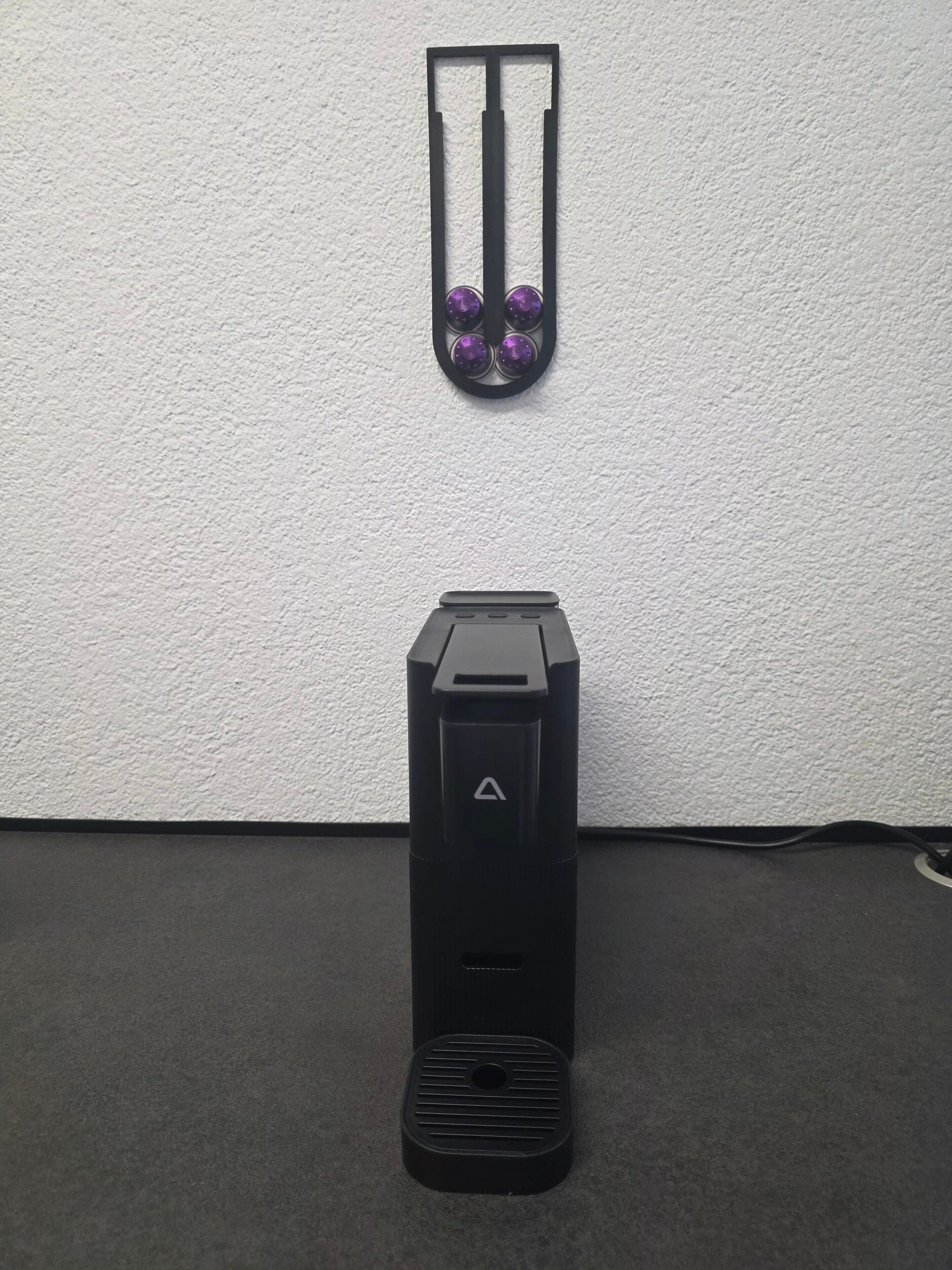 En svart elektronisk apparat med en triangulär logotyp på framsidan, placerad på en mörk yta mot en vit vägg.
