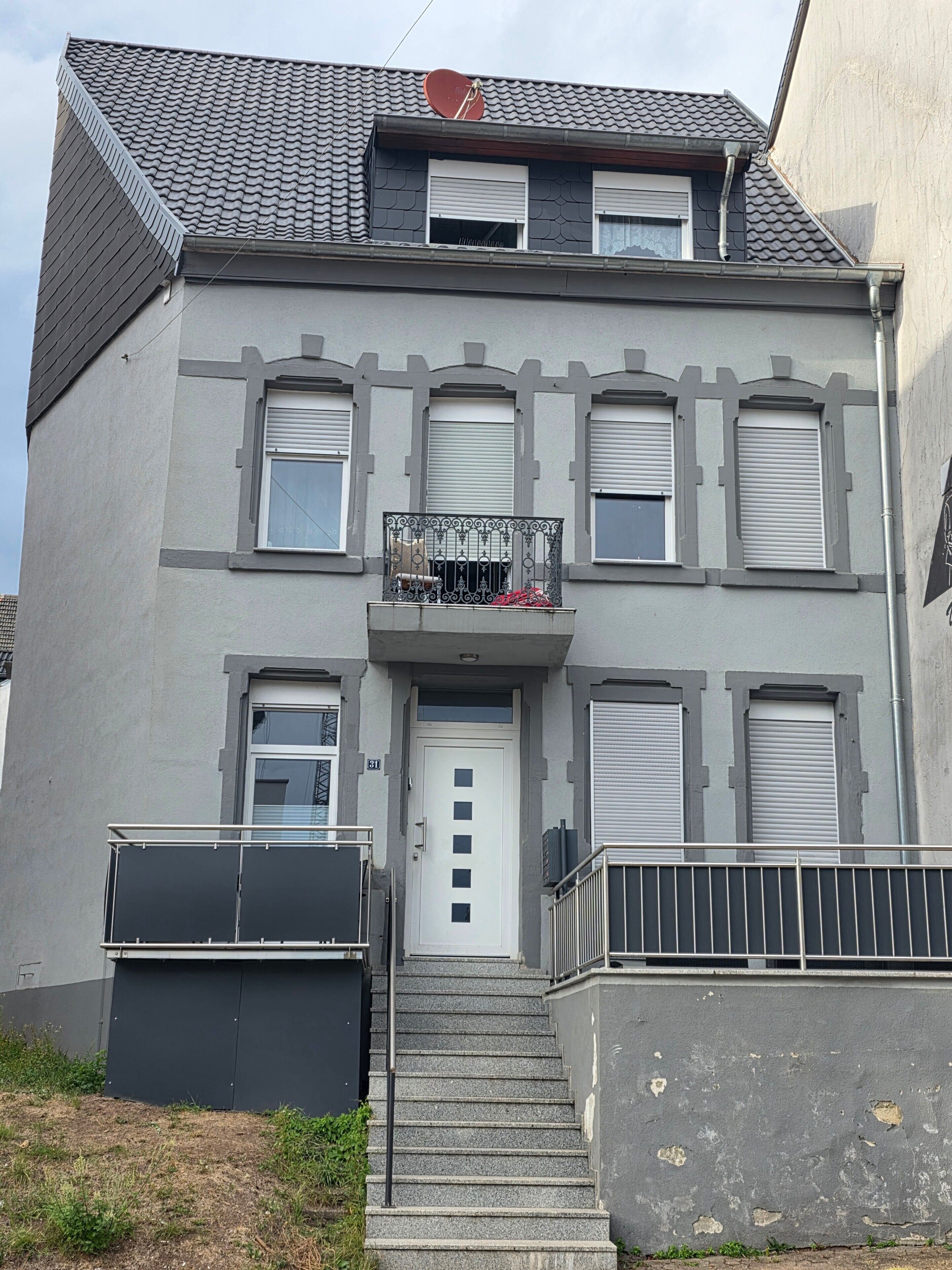 Ein zweistöckiges Gebäude mit einer grauen Fassade, einer weißen Tür und einem Balkon mit Metallgeländer.