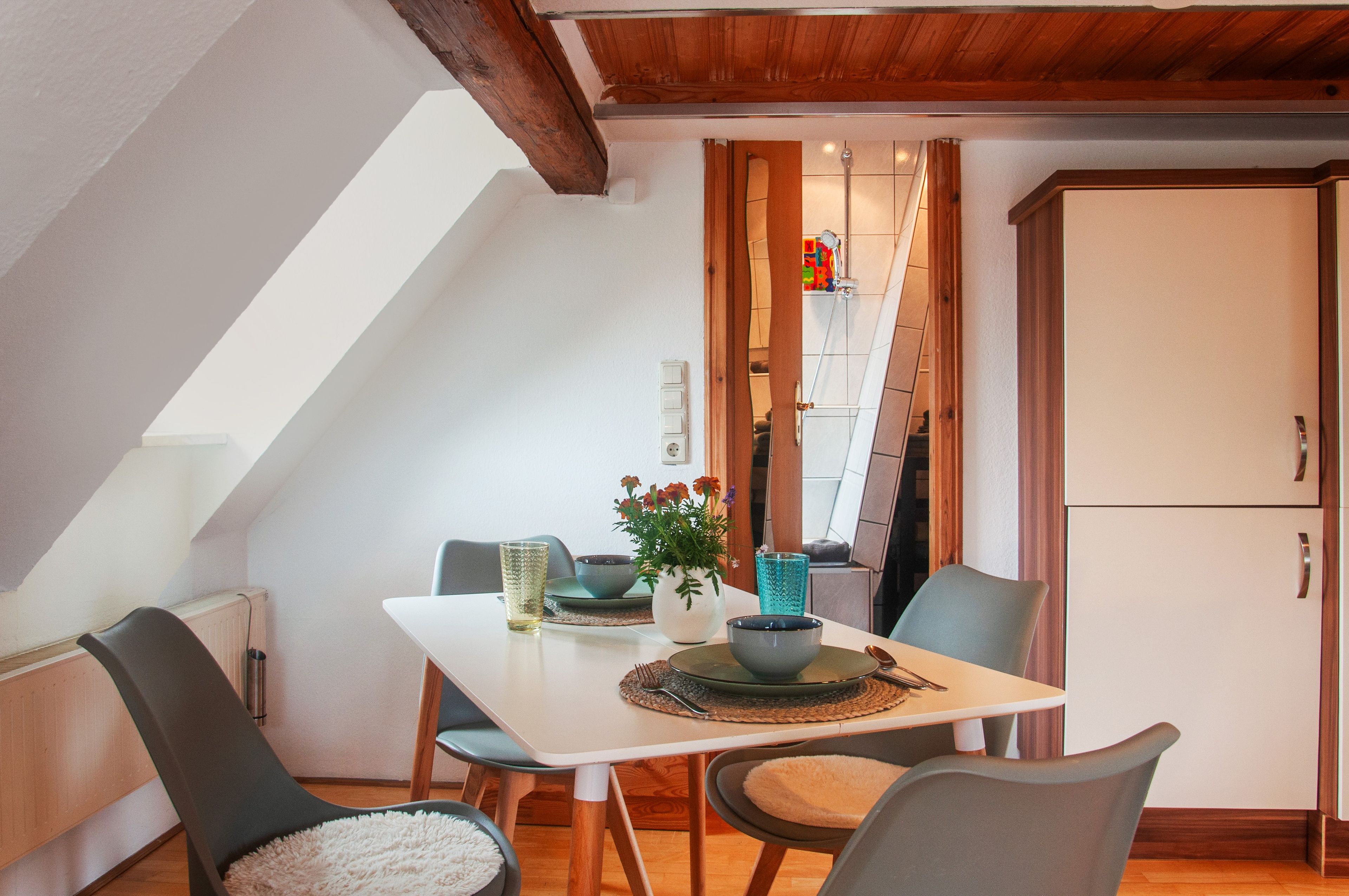 Een eetgedeelte met een witte tafel, grijze stoelen, een vaas met bloemen en een blauw glas.