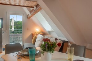 A dining area with a table set for two, a vase with flowers, and a view of a balcony with a railing and a chair.