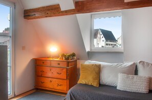 A bedroom with a wooden dresser, a bed with pillows, a window with a view of buildings, and a lamp on the dresser.