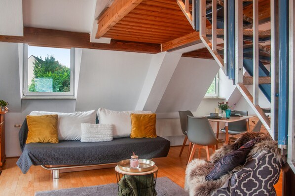 A loft-style living space with a sloped ceiling, a sofa with cushions, a dining area with a table and chairs, and a staircase.