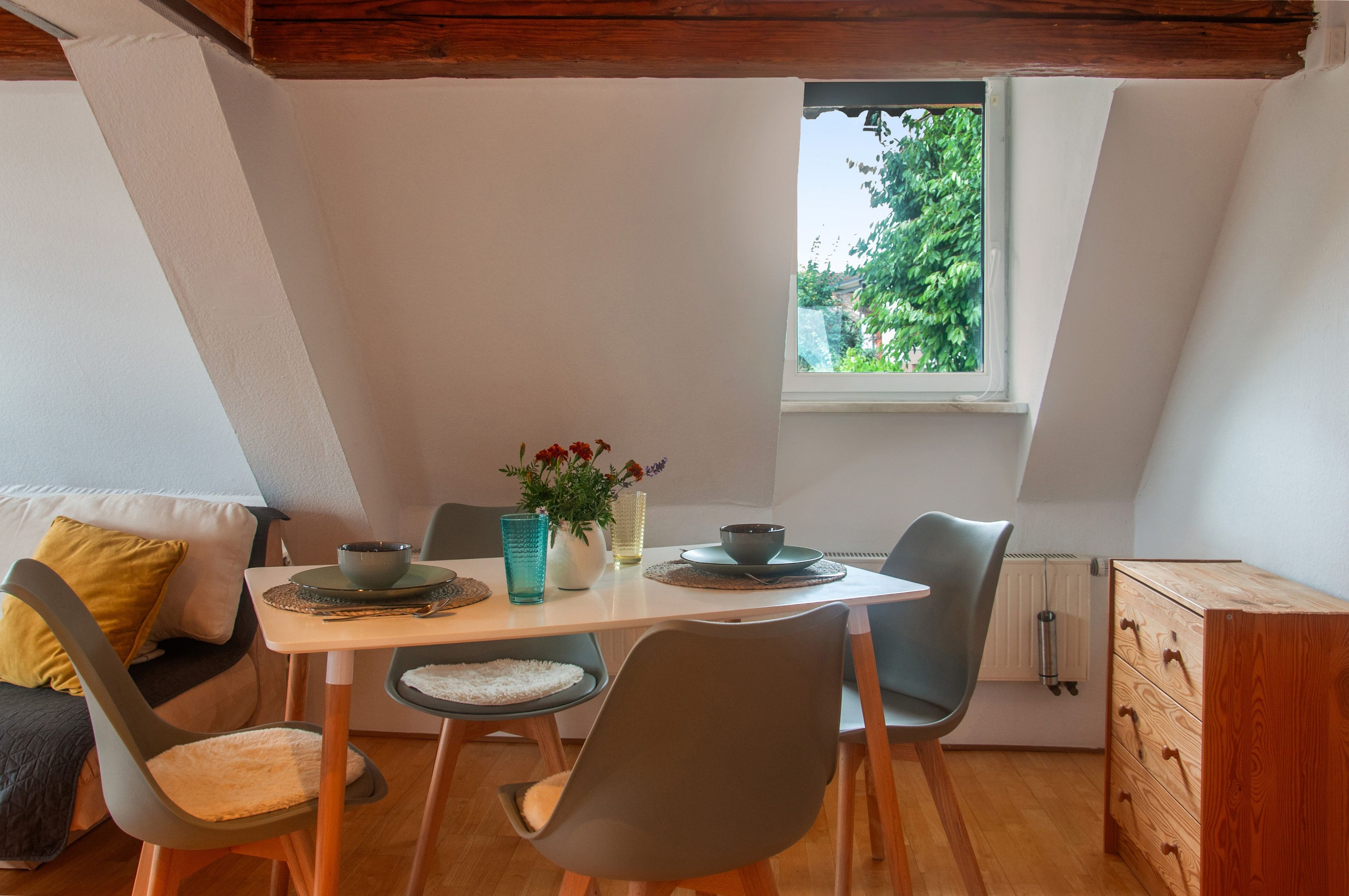 A dining area with a table set for two, chairs, a window with a view of greenery, and a radiator.