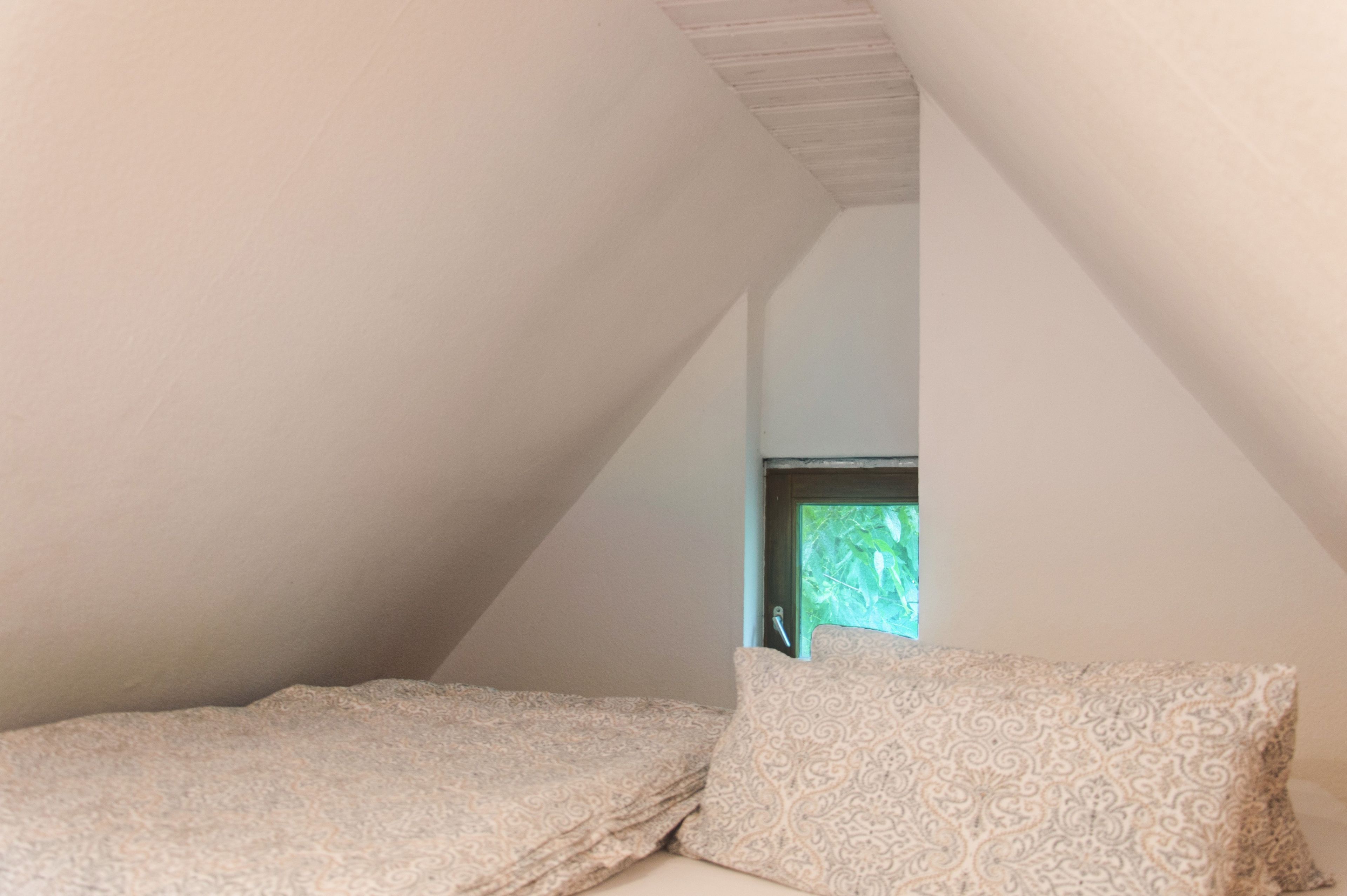 Un pequeño dormitorio abuhardillado con poca luz, techo inclinado, una ventana y una cama con ropa de cama estampada.