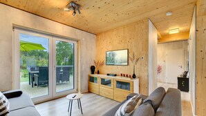 A modern living room with a wooden ceiling, a grey sofa, a small table, and a sliding glass door leading to an outdoor area. - Holiday Home for up to 6 Guests with Pool and Sauna (Freudenstadt)