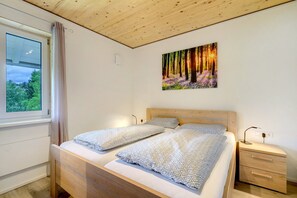 Ein Schlafzimmer mit Holzboden, einem Bett mit gemusterten Textilien, einem Nachttisch, einem Wandbild und einem Fenster mit Vorhängen.