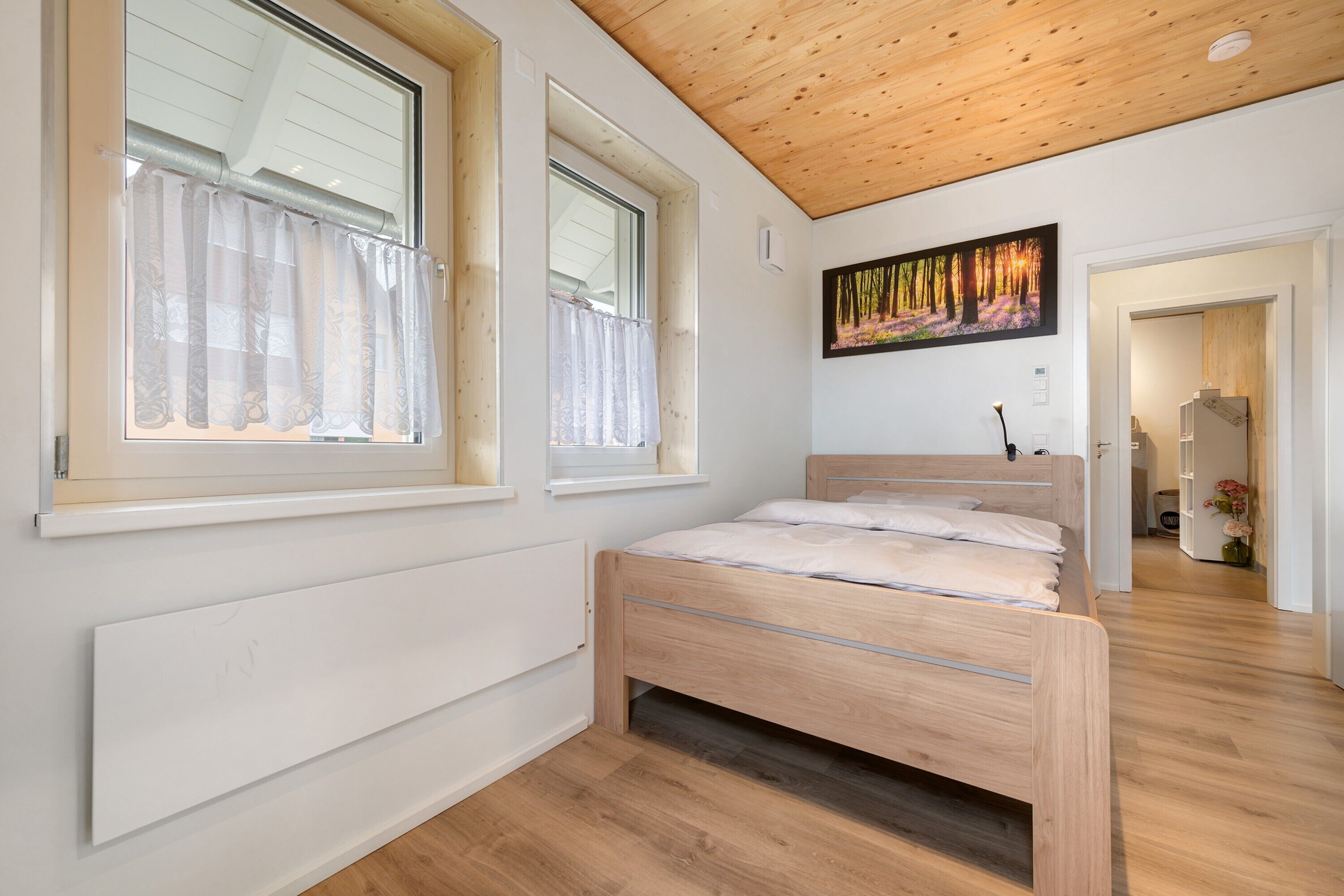 Um quarto com teto de madeira, uma cama com cabeceira, duas janelas com cortinas e um quadro na parede.