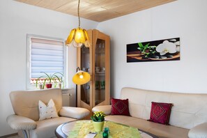 Un salon comprenant un canapé, des fauteuils, une table avec un vase, un meuble et une fenêtre avec des stores.