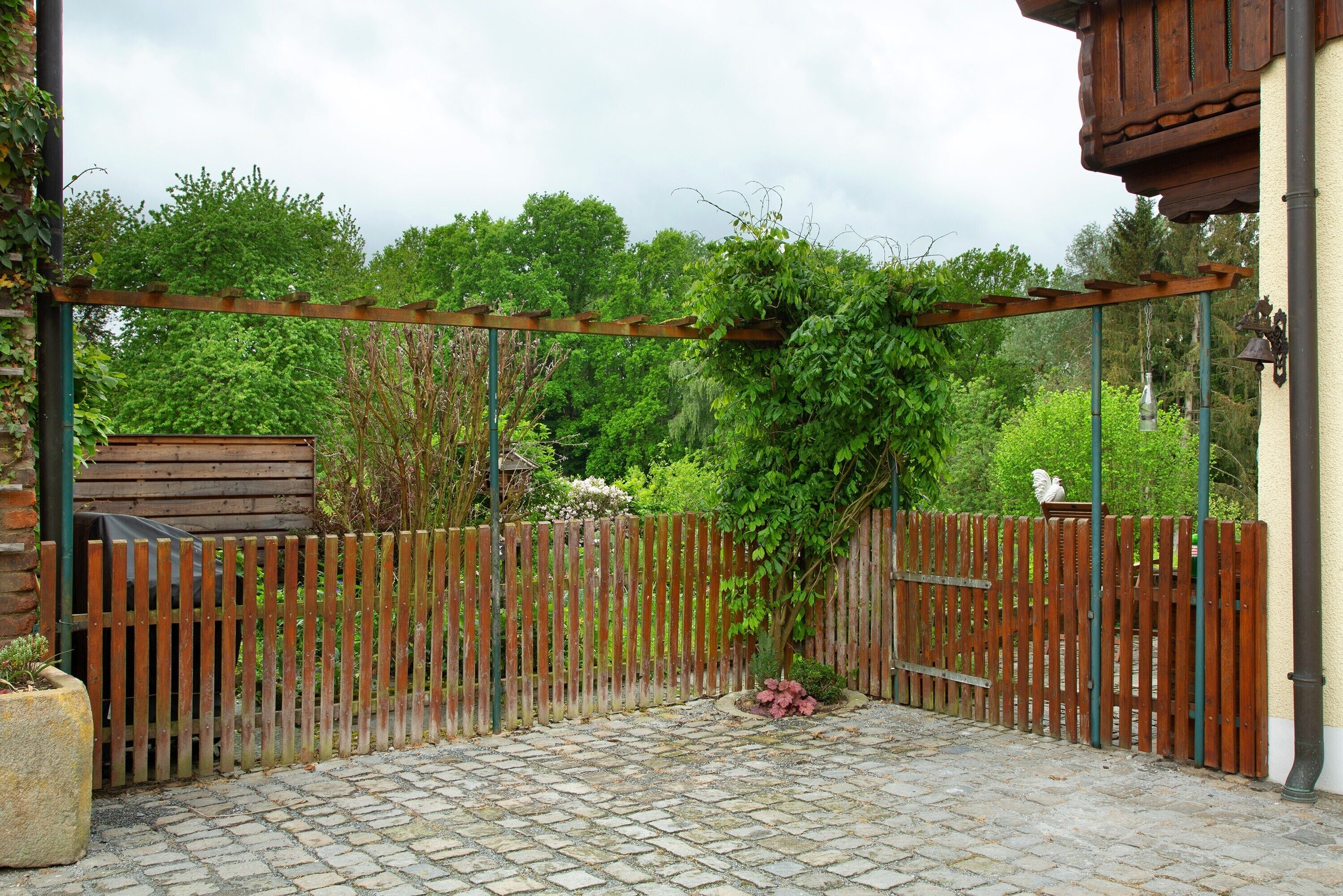 A garden patio with a wooden fence, a pergola, and a statue in the background.