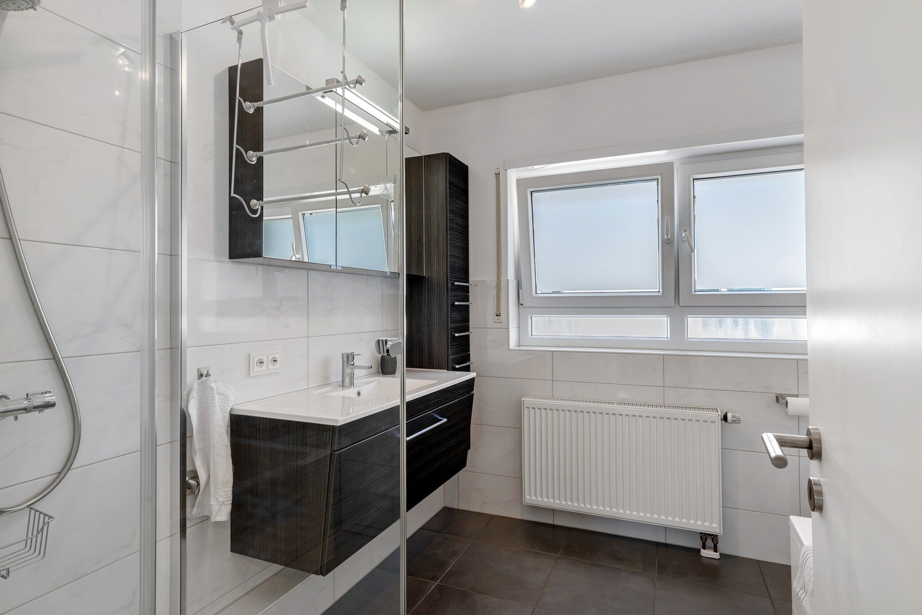 Ein modernes Badezimmer mit Duschkabine aus Glas, einem Waschtisch mit dunklem Unterschrank, einem weißen Waschbecken und einem Fenster für natürliches Licht.