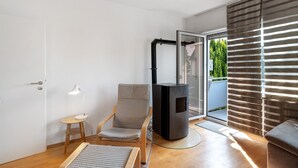 A modern living room with a wooden floor, a grey upholstered chair, a small wooden table with a lamp, a black fireplace, and a sliding glass door leading to a balcony with greenery.