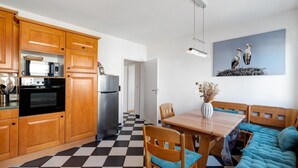 A kitchen with wooden cabinets, a microwave, and a refrigerator. There is a dining table with chairs and a sofa. A painting of storks is on the wall.