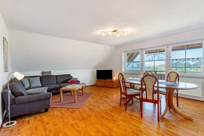 Una sala de estar con sofá, televisión, mesa de comedor, sillas y ventanas grandes.