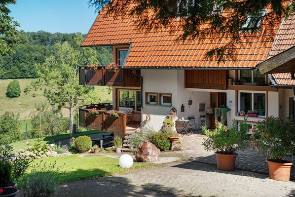 Une maison à deux étages, avec un toit recouvert de tuiles, un balcon orné de plantes suspendues et un jardin agrémenté de plantes en pot et d’un objet sphérique blanc.