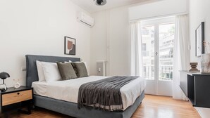 A modern bedroom with a grey bed, white walls, a radiator, and a large window with a view of buildings. - Acropolis Sunlight 2BR Aparment (Athina)