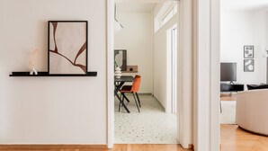 A modern living room with a wooden floor, a black and white abstract painting, a white vase with orange flowers, and a dining area with a table and chairs. - Acropolis Sunlight 2BR Aparment (Athina)