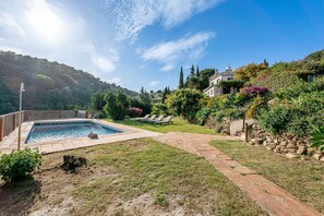 A swimming pool with a stone deck, a grassy area, and a hillside with trees and a house. - Brustar Villa del Sol Mijas (Mijas Costa)