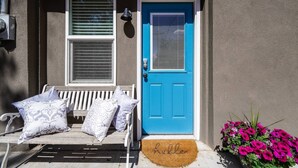 Una porta d'ingresso con una porta blu e un campanello bianco. C'è una panca con dei cuscini e un tappetino d'ingresso con la scritta "hello".