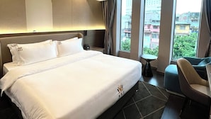 Una habitación de hotel moderna con una cama grande, un escritorio pequeño y vistas a la ciudad a través de amplias ventanas.