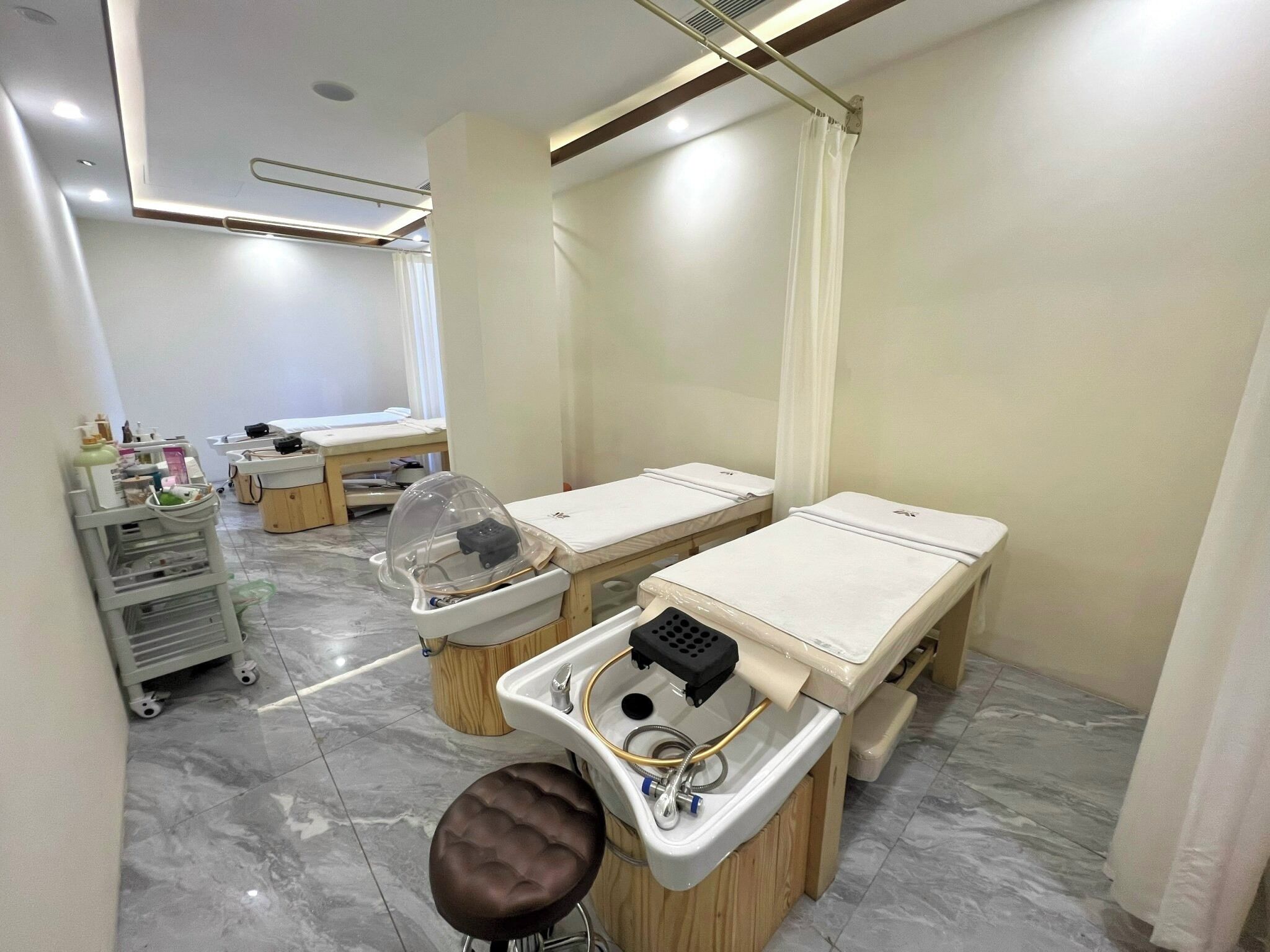 A clean, modern spa room with two treatment tables, a sink with a showerhead, and a stool.
