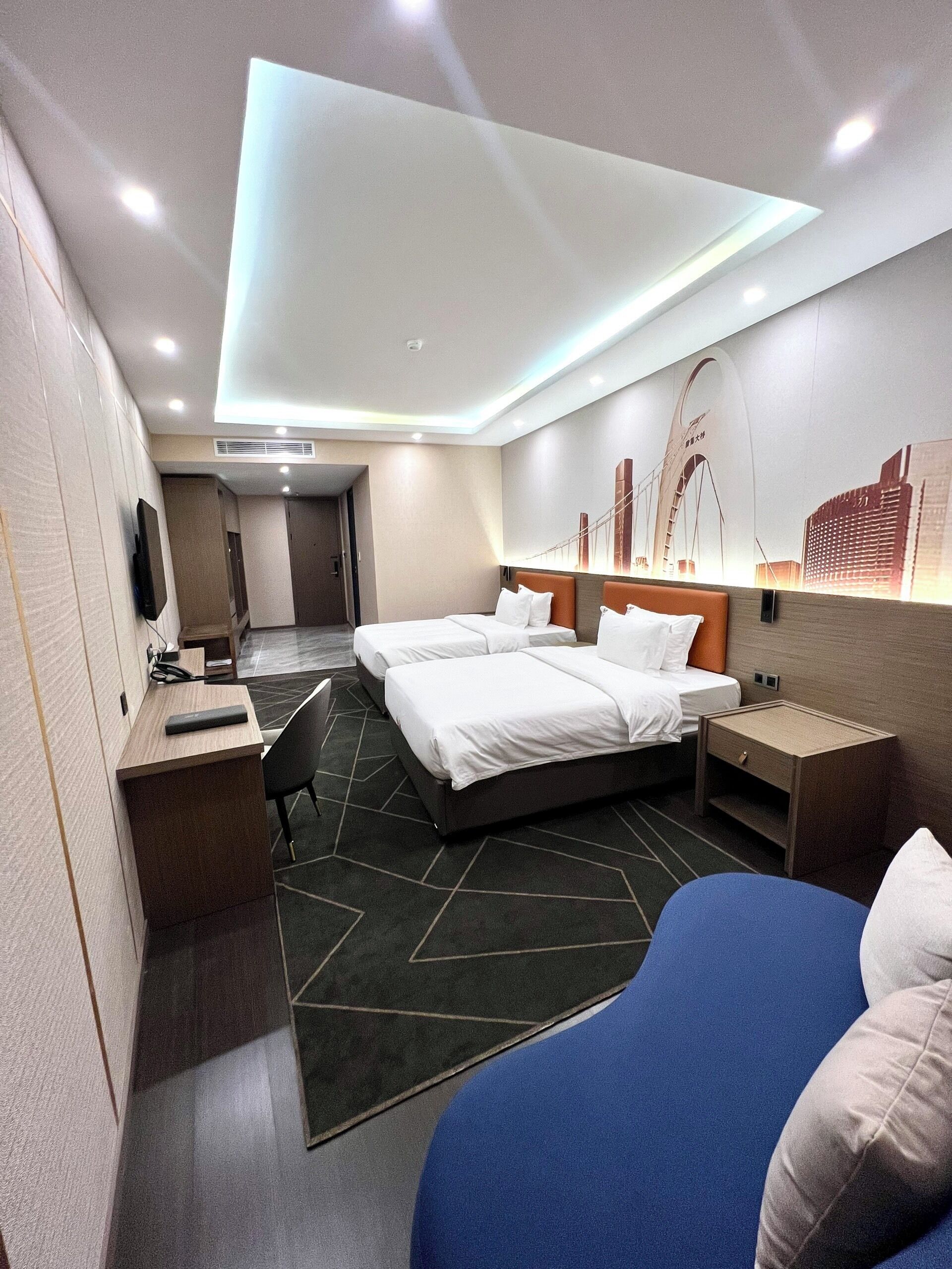 A modern hotel room with two beds, a desk, a chair, and a large window.