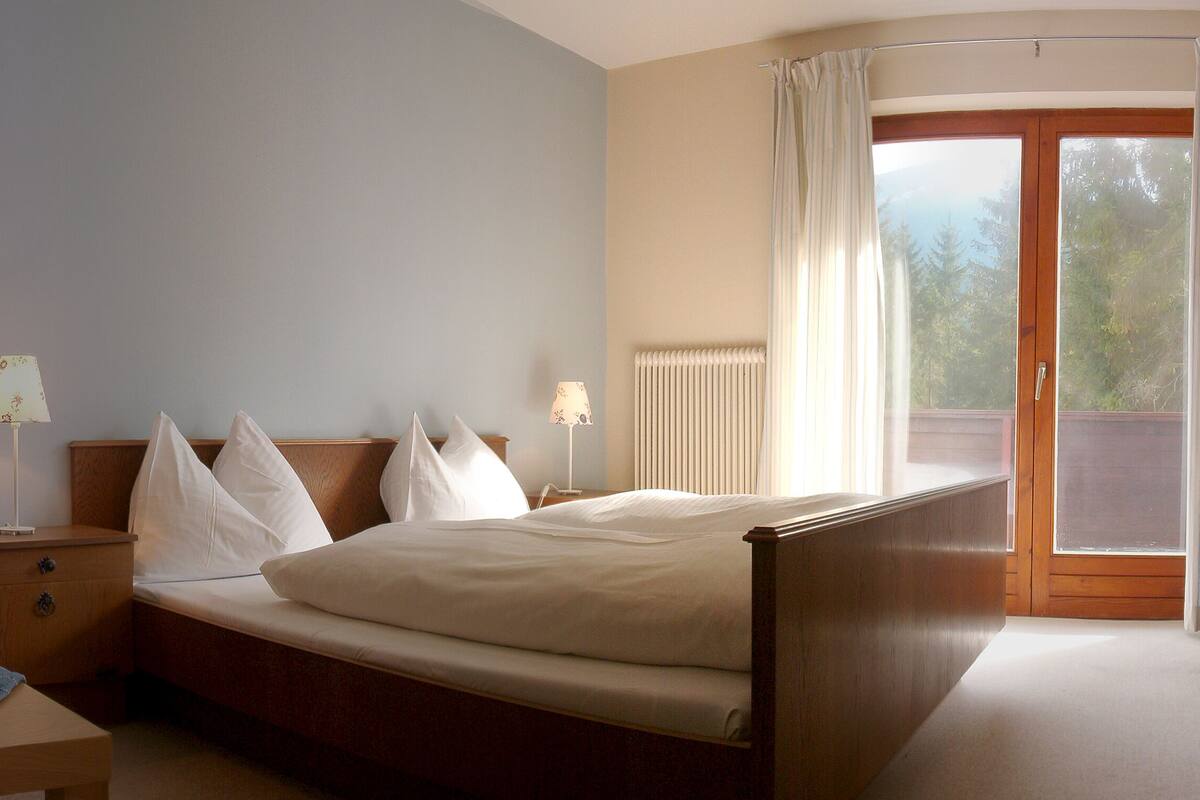 Un dormitorio con cama, mesitas de noche, una silla y vistas a través de un ventanal.