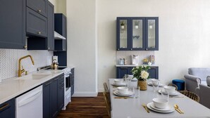 Una cocina moderna con armarios oscuros, encimera blanca y detalles dorados. Hay una zona de comedor con una mesa blanca dispuesta para cuatro personas, y un sofá al fondo.