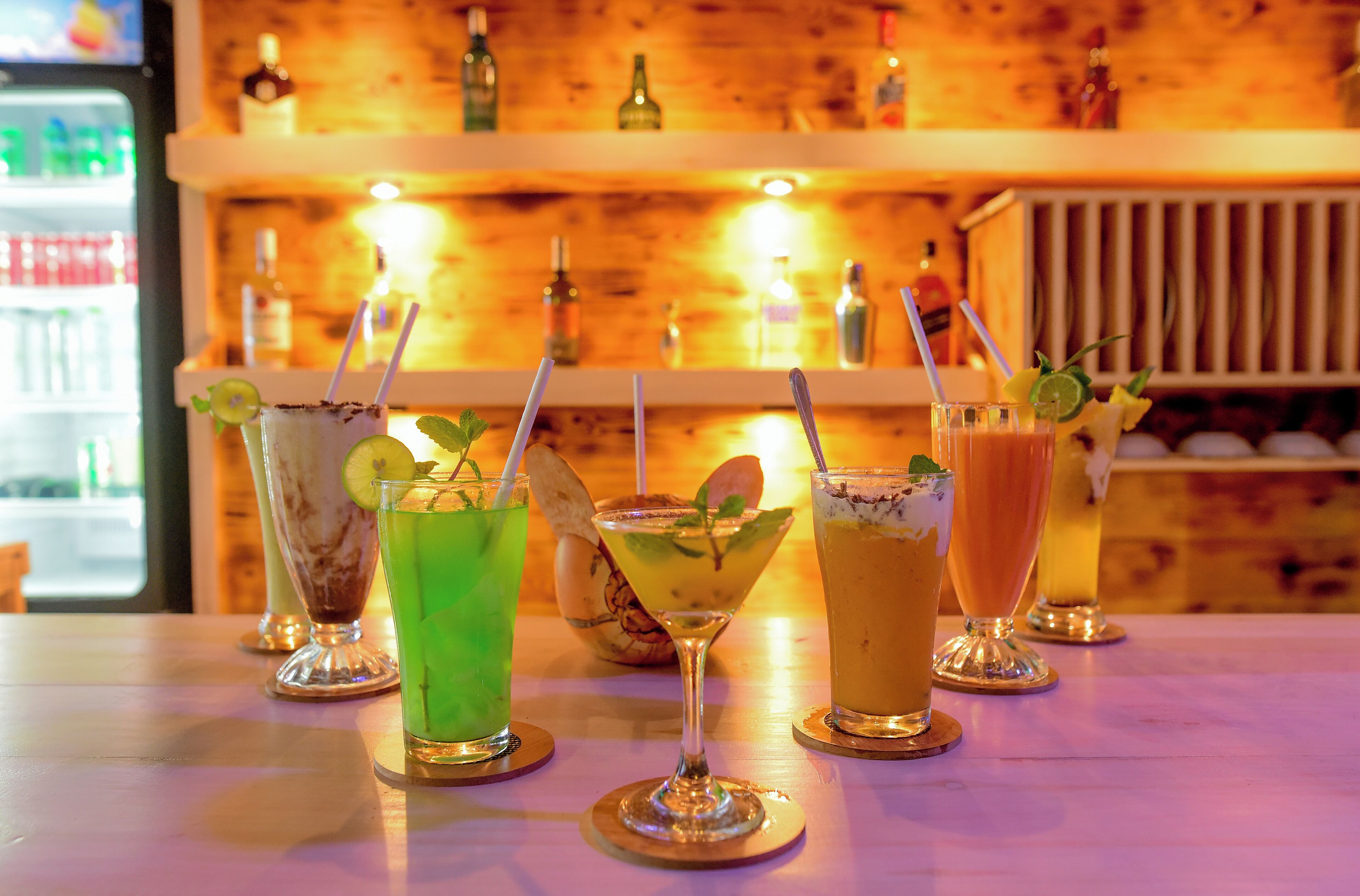 A bar with various drinks, including cocktails and a margarita, with a wooden backdrop and bottles of alcohol on shelves.