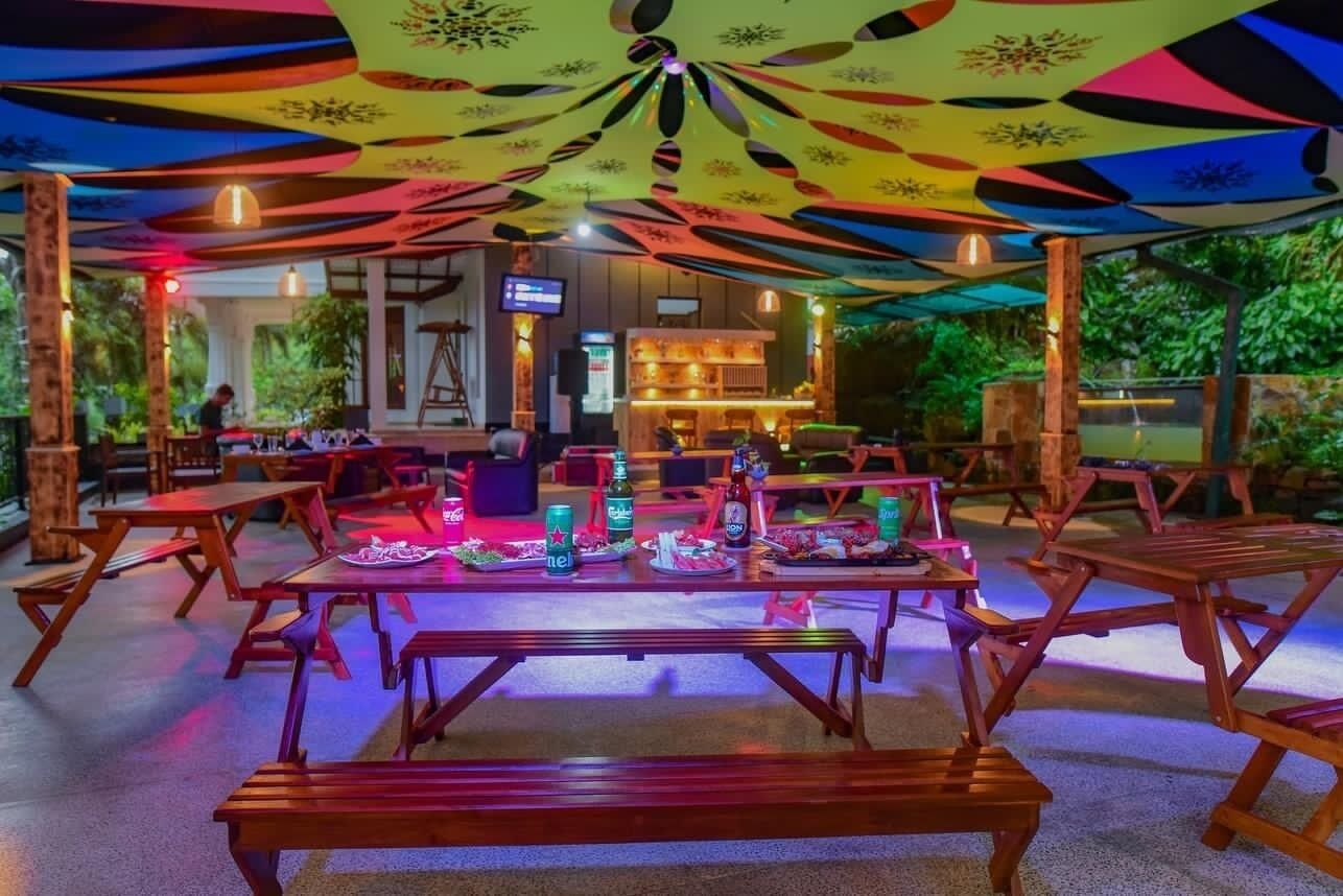 Un espace de restauration en plein air avec des tables et des bancs en bois, un bar en arrière-plan et un plafond aux couleurs vives.