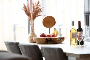 A dining table with a wooden bowl containing apples and bananas, a vase with pampas grass, and two wine bottles with glasses. - Holiday Home in Zevenhuizen with Sauna (Zevenhuizen)