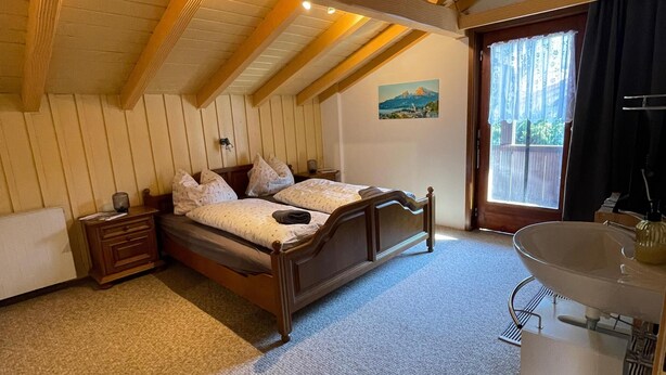 Une chambre avec un lit en bois, une table de chevet, une commode, une fenêtre donnant sur un beau paysage, et une salle de bain avec un lavabo et un miroir.