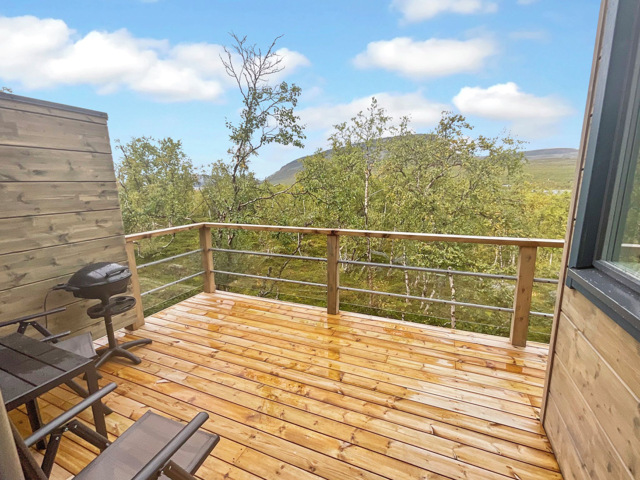 Eine Holzterrasse mit Metallgeländer, einem Grill und einem Tisch mit Stühlen.