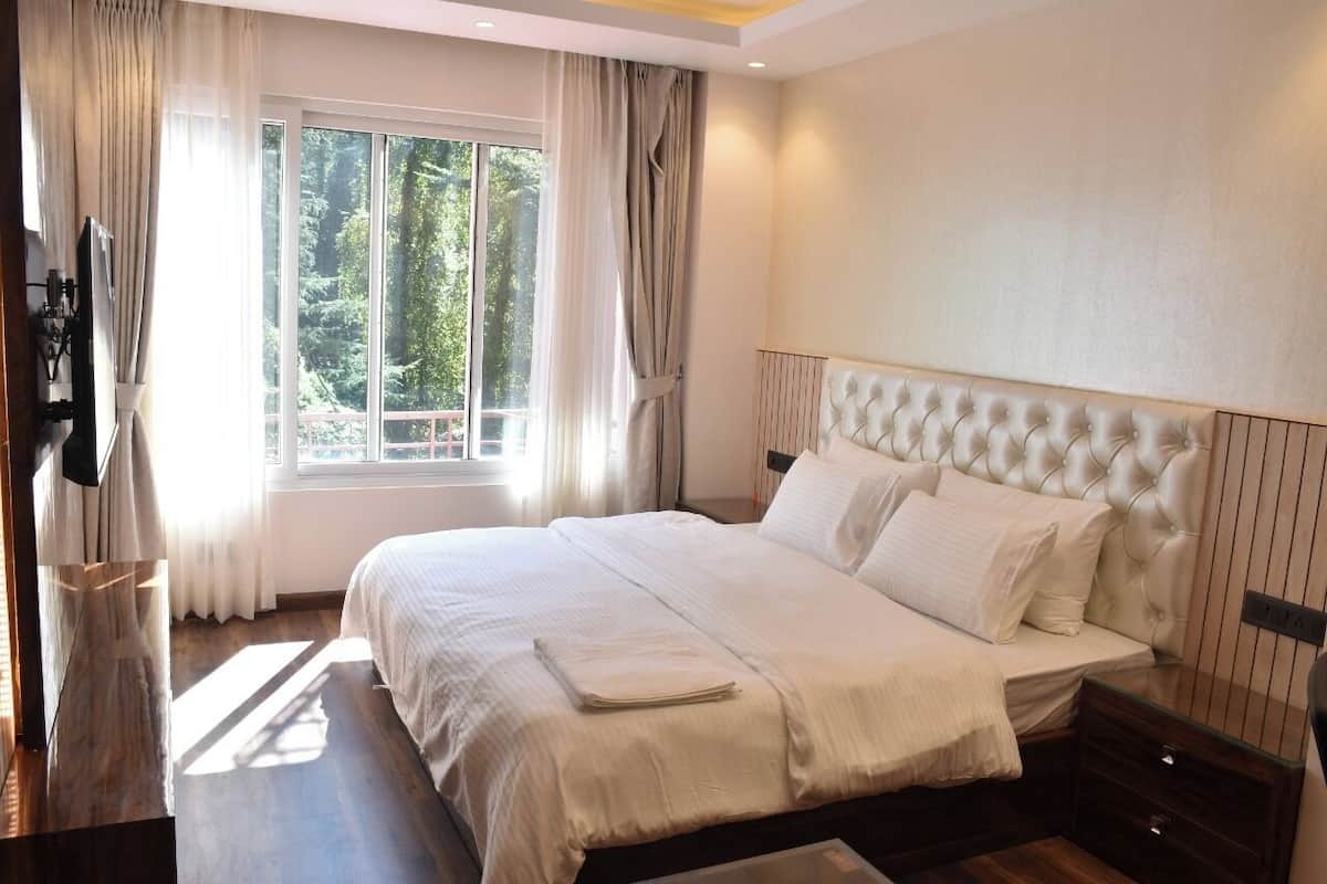 A bedroom with a large bed, a window with curtains, a wooden floor, and a wall-mounted TV.