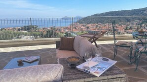 Una terraza con mobiliario de mimbre, una mesa con un cuenco de frutos secos, una copa de vino y una revista. Se aprecia una vista panorámica de un pueblo costero y colinas al fondo.